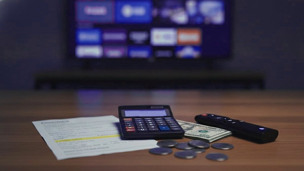 Cover for “How much does IPTV cost? A buyer’s guide to terms, screens and extras”: a calculator with a blank display, papers, banknotes, coins and a remote on a wooden table in front of a television