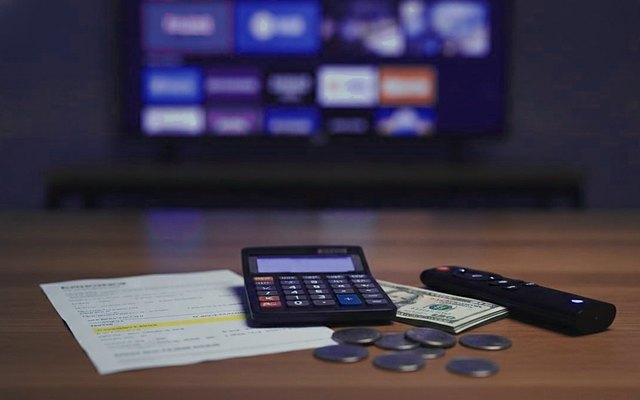 Cover for “How much does IPTV cost? A buyer’s guide to terms, screens and extras”: a calculator with a blank display, papers, banknotes, coins and a remote on a wooden table in front of a television
